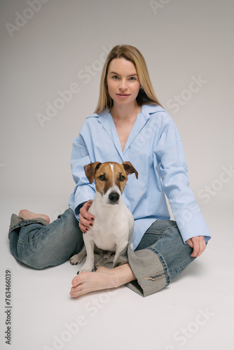 Woman and dog happy friends sitting on the floor looking at camera and smiling. Professional photo studio. Pretty blonde woman in blue jeans shirt and pet Jack Russell terrier. Vertical composition