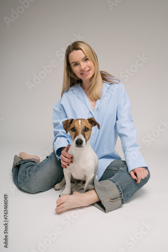 Woman and dog friends sitting on the floor looking at camera and smiling. Professional photo studio. Pretty blonde woman in blue clothes and pet Jack Russell terrier. Vertical composition