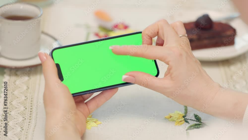 Close-up Of Mobile Phone With Green Mock-up Screen In Female Hands. Woman Uses Smartphone While Resting In Cafe, Drinks Tea and Eats Dessert Close-up Of Mobile Phone With Green Mock-up Screen In Female Hands. Woman Uses Smartphone While Resting In Cafe, Drinks Tea and Eats Dessert