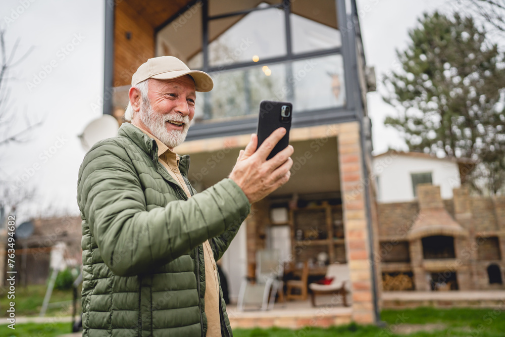 One senior man stand in front of tiny house in day use smart phone One senior man stand in front of tiny house in day use smart phone