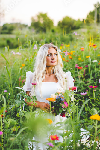 Beautiful Blonde Model Sitting in Zinnia Wildflower Garden, Colorful Golden Hour Sunset Bridal Portrait of Bridal Model with Loose Hair Wearing Modern Wedding Dress with Puffy Sleeves photo