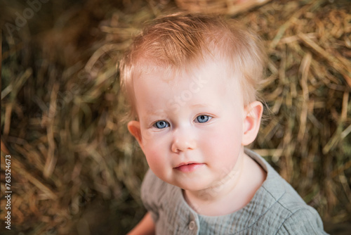 A baby with blue eyes and blonde hair is sitting in a hay bale.