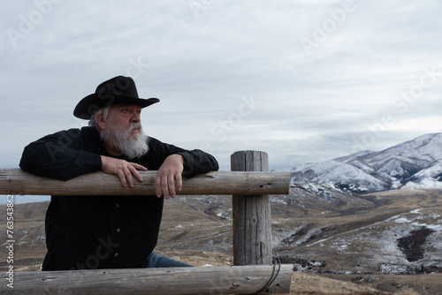 Slika na platnu Elderly Caucasian male rancher man with cowboy hat leaning on wooden fence looki