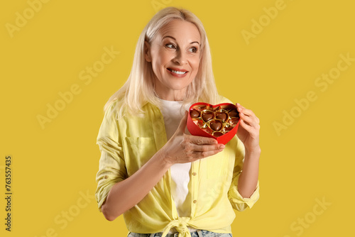 Mature woman with box of heart-shaped chocolate candies on yellow background