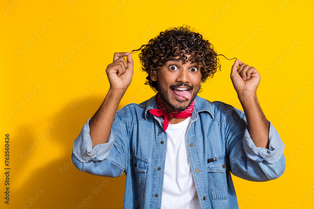 Photo portrait of nice young male hold curly strands stick tongue wear trendy denim outfit red scarf isolated on yellow color background Photo portrait of nice young male hold curly strands stick tongue wear trendy denim outfit red scarf isolated on yellow color background