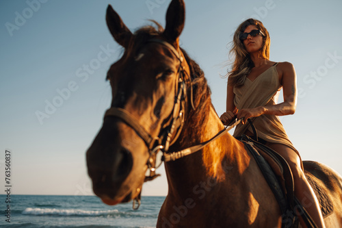 A charming young woman is riding a horse on the ocean beach at sunset.
