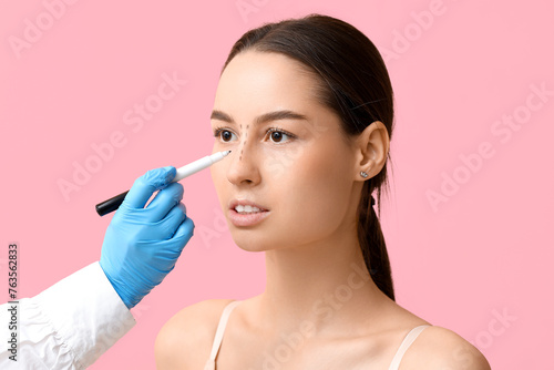 Doctor in protective gloves drawing marks on woman s nose for plastic surgery against pink background