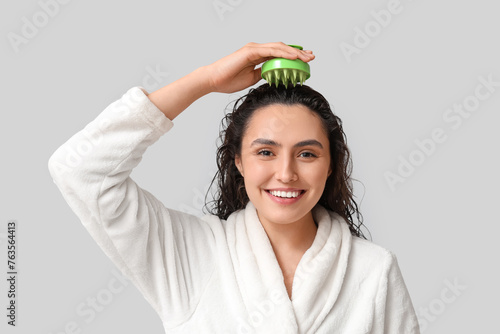 Beautiful young woman using hair scalp massager on grey background