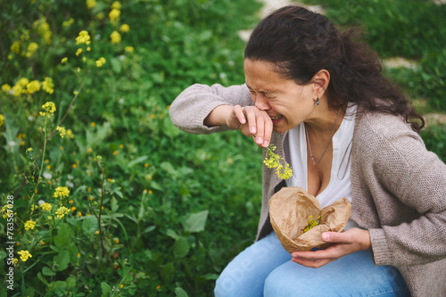 A young woman sneezing while gathering wildflowers in the meadow outdoors, suffering from allergy in the spring time photo