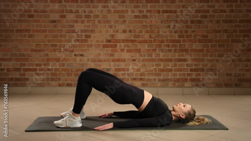 Performing hip bridges on a mat, the dedicated trainer is targeting glute muscles and lower back, promoting better posture and core stability in a serene gym setting with a brick wall background.