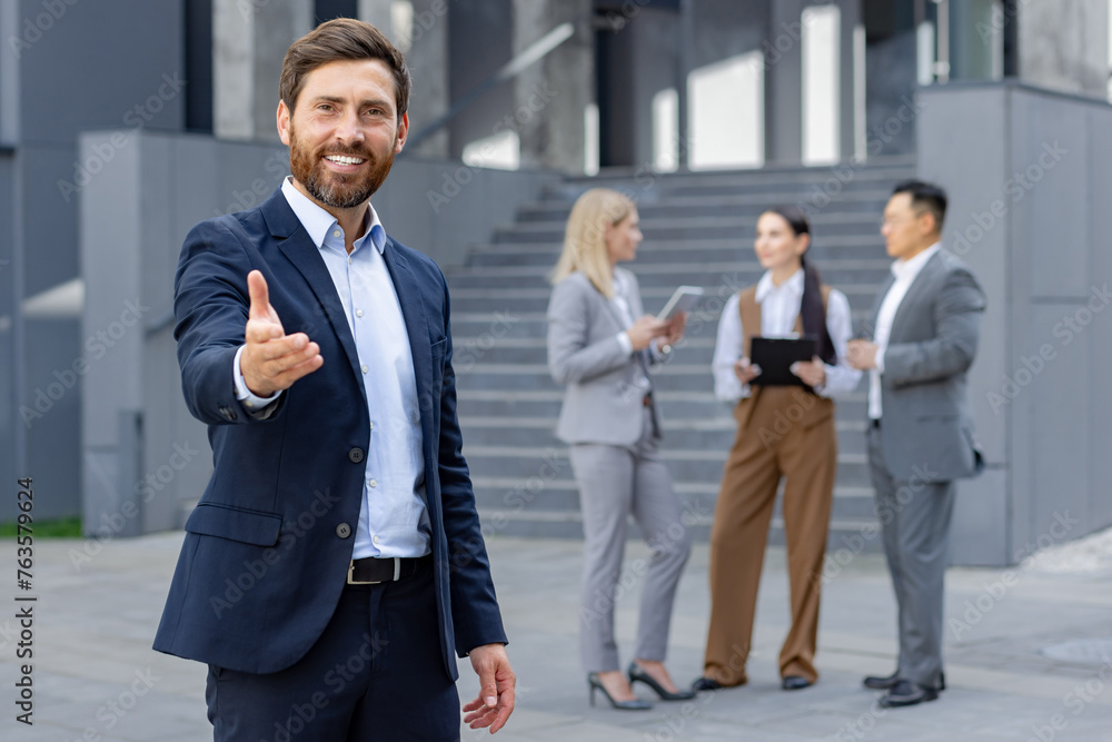 A professional businessman extending his hand for a handshake in front of colleagues outside a modern office building. A professional businessman extending his hand for a handshake in front of colleagues outside a modern office building.