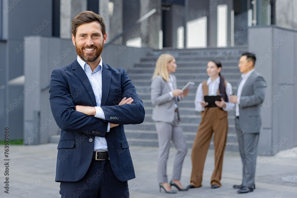 A well-dressed businessman stands confidently outdoors with his colleagues discussing in the background. A well-dressed businessman stands confidently outdoors with his colleagues discussing in the background.