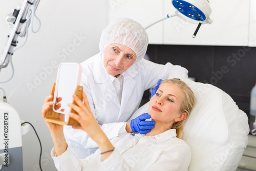 Woman looking in mirror during doctor s appointment in cosmetological clinic. Elderly female cosmetologist standing beside.