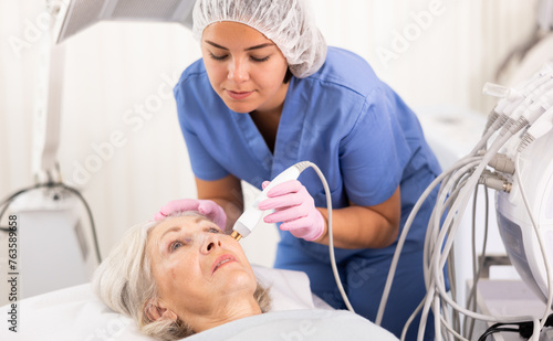 Elderly woman receiving non-surgical rejuvenation procedure from female cosmetologist  RF face lifting with use of electric device equipment in spa center