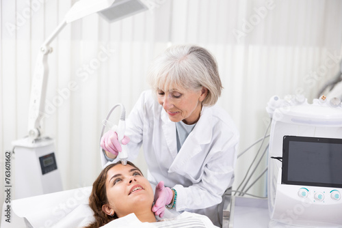 Careful old woman doctor conducting vacuum cleaning of young woman patient's face in aesthetic clinic