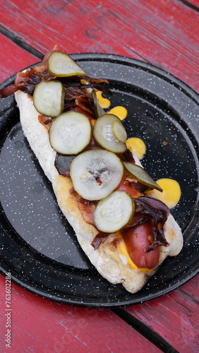American food. Fusion. Closeup view of a gourmet hot dog with crispy bacon, cheddar cheese and cucumber slices, presente don a black dish on the red wooden table.