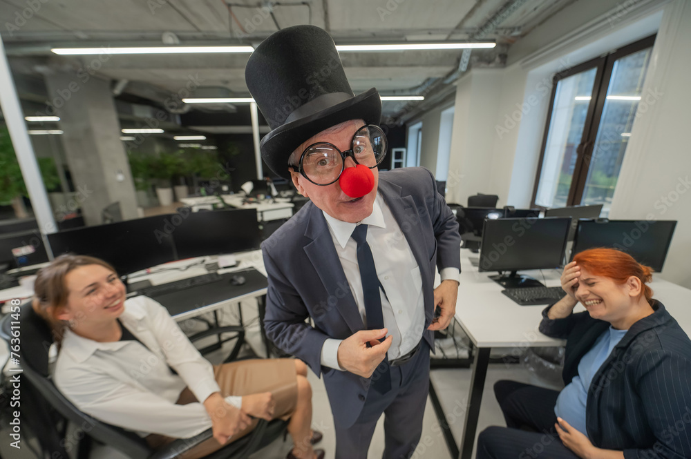 An elderly Caucasian man in a clown costume amuses two Caucasian women in the office. An elderly Caucasian man in a clown costume amuses two Caucasian women in the office.