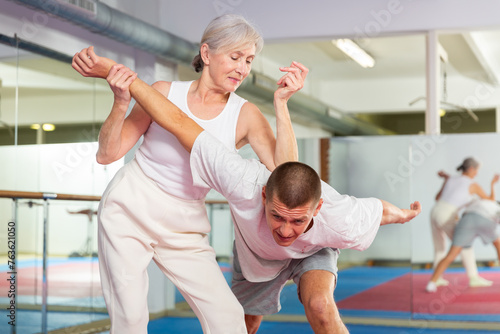 Focused elderly woman performing elbow strike and wristlock  painful control move to immobilize male opponent during self defense training in gym