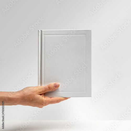 Female hands holding a blank notebook from below. Hardcover book. Matte white. Blank book mockup. Textured paper. (real photo) 3D render.  (ID: 763632620)