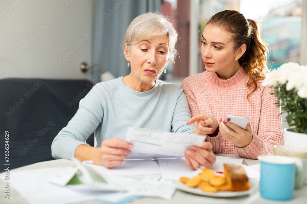 Interested focused elderly woman sitting in dining room with adult daughter using smartphone and checking utility bills.