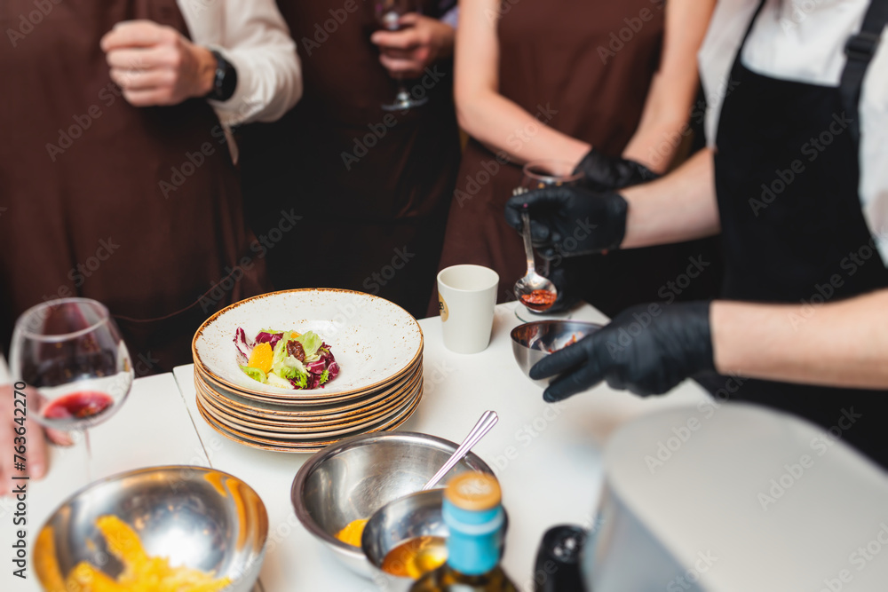 Group of guests in a cooking class studio, adults preparing different dishes in the kitchen together, people in aprons learn on culinary master class, chef uniform, hands in gloves, italian cuisine Group of guests in a cooking class studio, adults preparing different dishes in the kitchen together, people in aprons learn on culinary master class, chef uniform, hands in gloves, italian cuisine