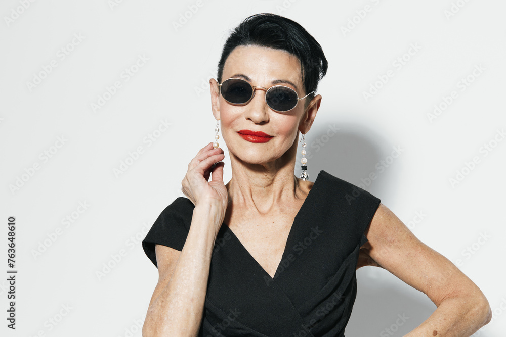 Elegant senior woman in sunglasses and black dress posing in front of white wall for camera portrait Elegant senior woman in sunglasses and black dress posing in front of white wall for camera portrait
