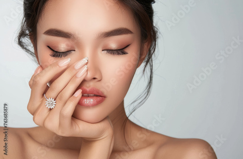Beautiful asian woman with perfect manicure and makeup posing for nail art studio, eyes closed, hand covering half of her face, holding a diamond ring on her finger