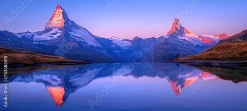 In the Swiss Alps, two majestic mountains stand tall against a clear blue sky at sunrise. The peaks reflect in the still waters of an alpine lake