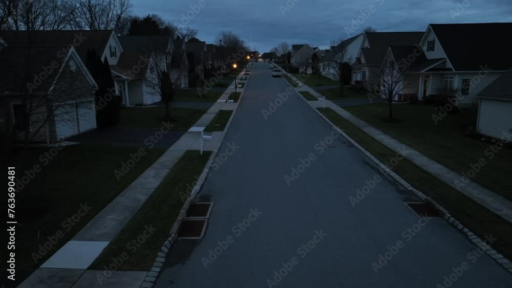 Aerial view of a symmetrical American suburb at twilight, with streetlights on, showcasing identical houses and bare trees. Low drone truck shot at dusk.