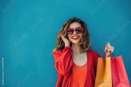 Photo of happy woman holding shopping bags isolated on blue background, wearing sunglasses and having fun during a sale at a store or mall