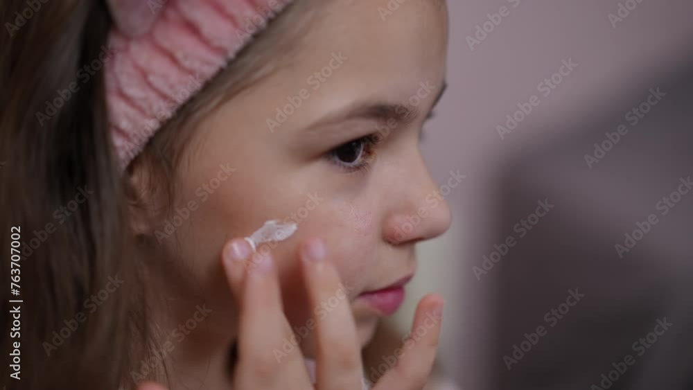 Slow motion. Close-up. The little fashionista smiles, as she applies white cream to her face with her finger and gently rubs it with her hands Slow motion. Close-up. The little fashionista smiles, as she applies white cream to her face with her finger and gently rubs it with her hands