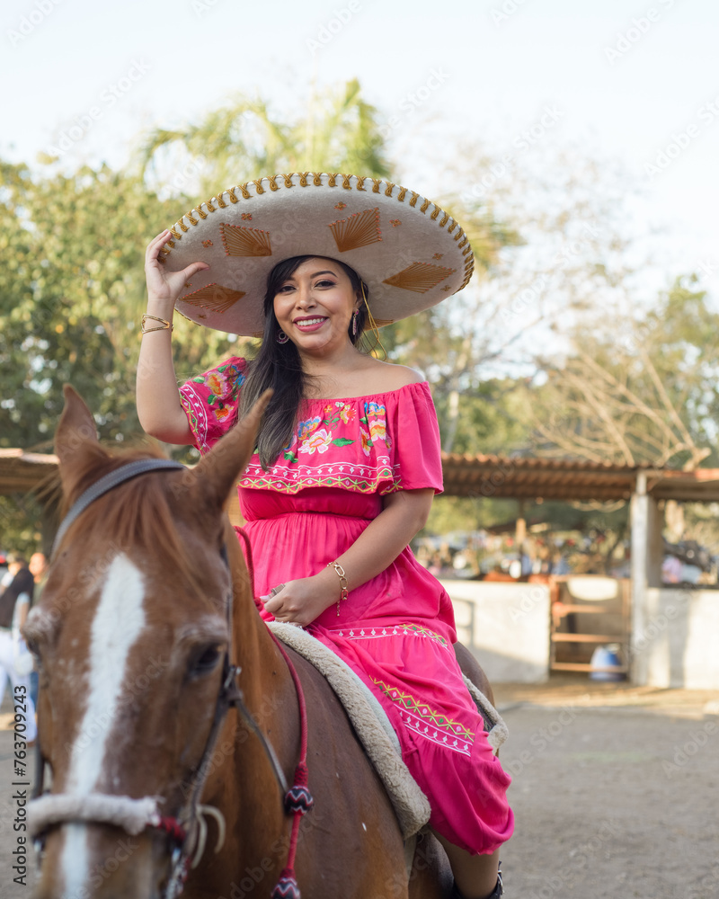 Obraz premium Mexican woman wearing traditional dress and charro hat on horseback. Cinco de Mayo celebration.