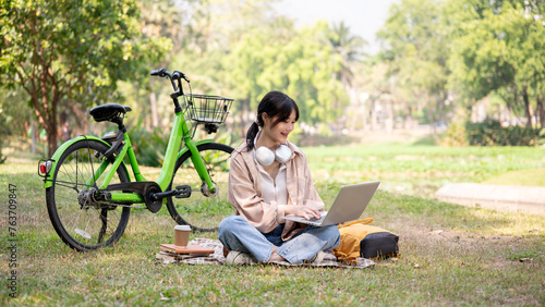 A happy young Asian woman is working remotely in a green park, using her laptop computer.