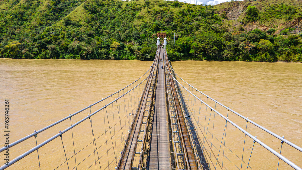 Fototapeta premium Western Bridge in Santa Fe de Antioquia, Colombia, spanning a muddy river with lush green hills in the background