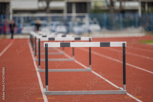 Obraz na płótnie Hurdles for athletic running standing in a row. Open space on stadium, running track with a row of hurdles. Sunny day, person in bokeh in the background w salonie