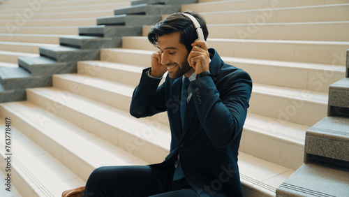 Project manager listening and enjoy music from headphone while sitting at stairs. Professional businessman wearing suit outfit while dancing and moving to music. Happy man listen funny song. Exultant.