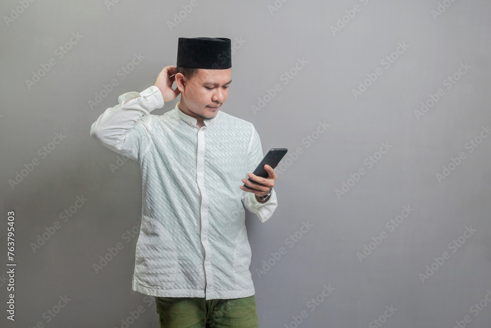 Asian Muslim man standing scratching his head and looking the smartphone Asian Muslim man standing scratching his head and looking the smartphone