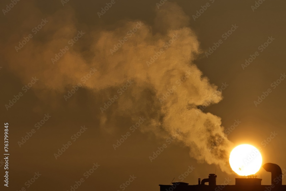 Smoke coming out of the chimney of a multi-family building against the background of the rising sun, air pollution, heating of buildings with gas and coal furnaces, fossil fuels.
