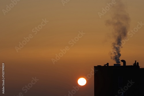 Smoke coming out of the chimney of a multi-family building against the background of the rising sun, air pollution, heating of buildings with gas and coal furnaces, fossil fuels.