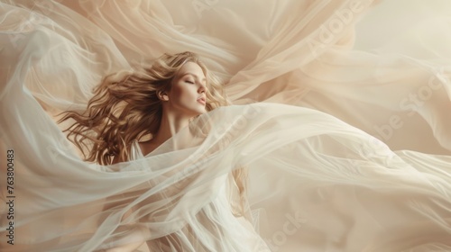 Beautiful young woman wrapped in transparent fabric. Fluttering veil on a beige background