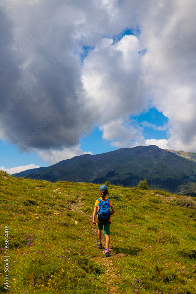hiking in the mountains. A little boy with a backpack walks along a path against a backdrop of mountains with his back to the camera.