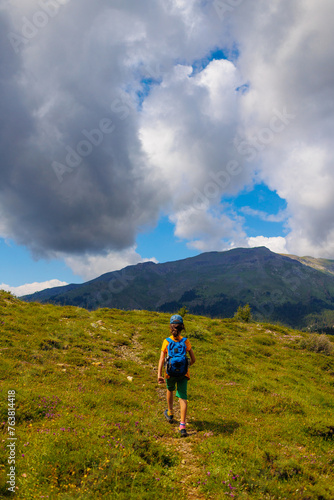 hiking in the mountains. A little boy with a backpack walks along a path against a backdrop of mountains with his back to the camera.