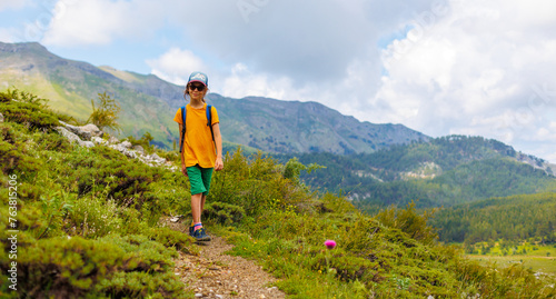 hiking in the mountains. A little boy with a backpack walks along a path against a background of mountains and clouds. Active healthy lifestyle on weekend hike journey.