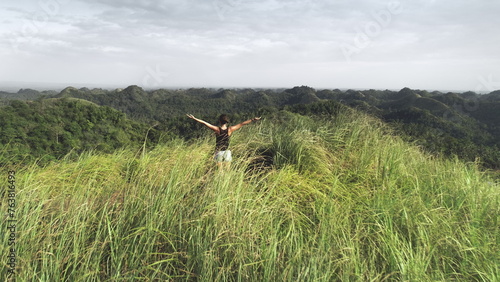 Aerial mountain top: woman rise-up hands at high grass peak of Philippines hills, Asia. Epic landscape with back view girl. Majestic Asian nature with tourist admire. Cinematic freedom drone shot