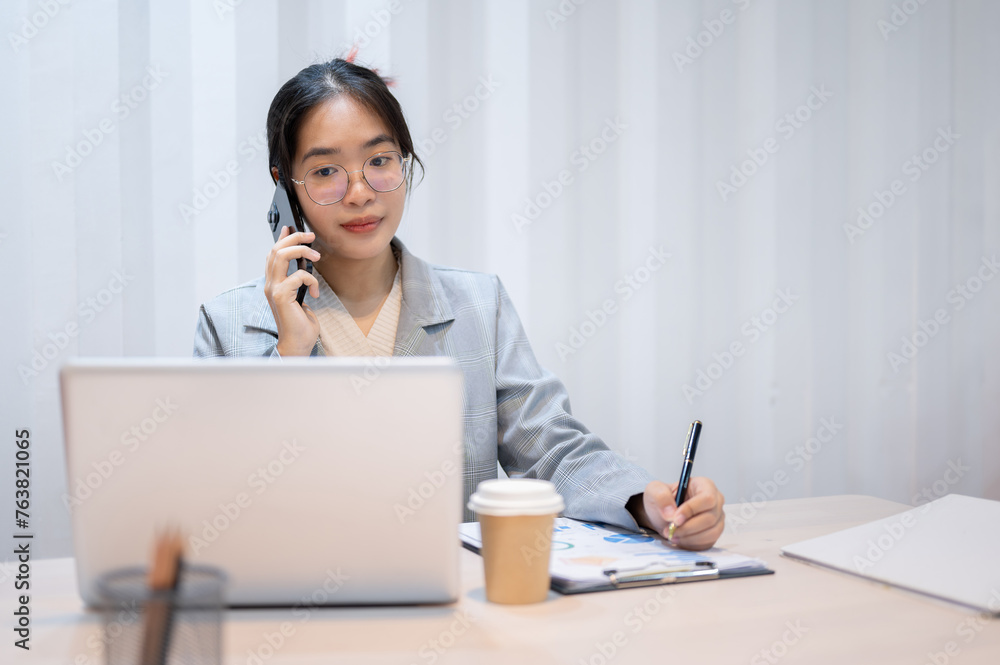 A businesswoman is talking on the phone, taking notes, and reading information on her laptop. A businesswoman is talking on the phone, taking notes, and reading information on her laptop.