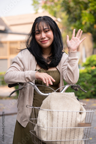 A smiling woman looking and waving her hand at the camera while traveling in the city on her bike. © bongkarn