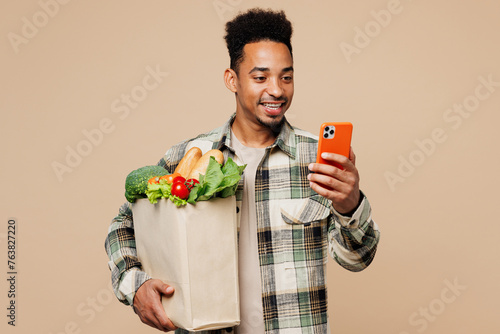 Young smiling man wear grey shirt hold paper bag for takeaway mock up with food products use mobile cell phone isolated on plain pastel light beige background Delivery service from shop or restaurant