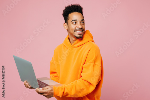 Side view young IT man of African American ethnicity wear yellow hoody casual clothes hold use work on laptop pc computer look aside isolated on plain pastel pink background studio. Lifestyle concept