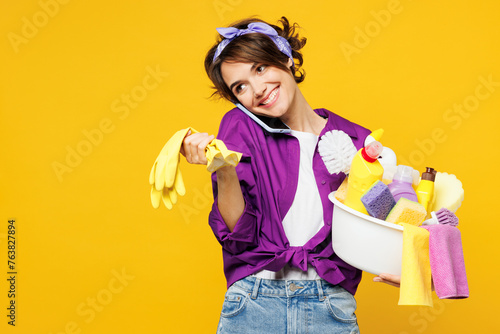 Young smiling woman wears purple shirt hold basin with detergent bottles do housework tidy up talk speak on mobile cell phone isolated on plain yellow background studio portrait. Housekeeping concept.