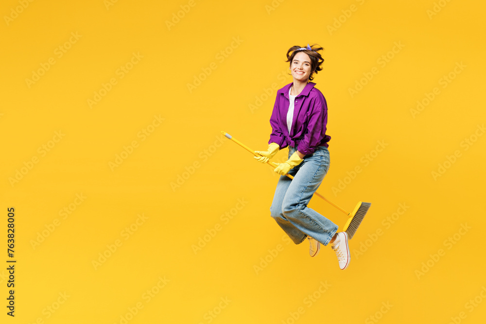 Full body young woman wears purple shirt casual clothes do housework tidy up jump high hold brush broom jump high pov fly like witch isolated on plain yellow background studio. Housekeeping concept.